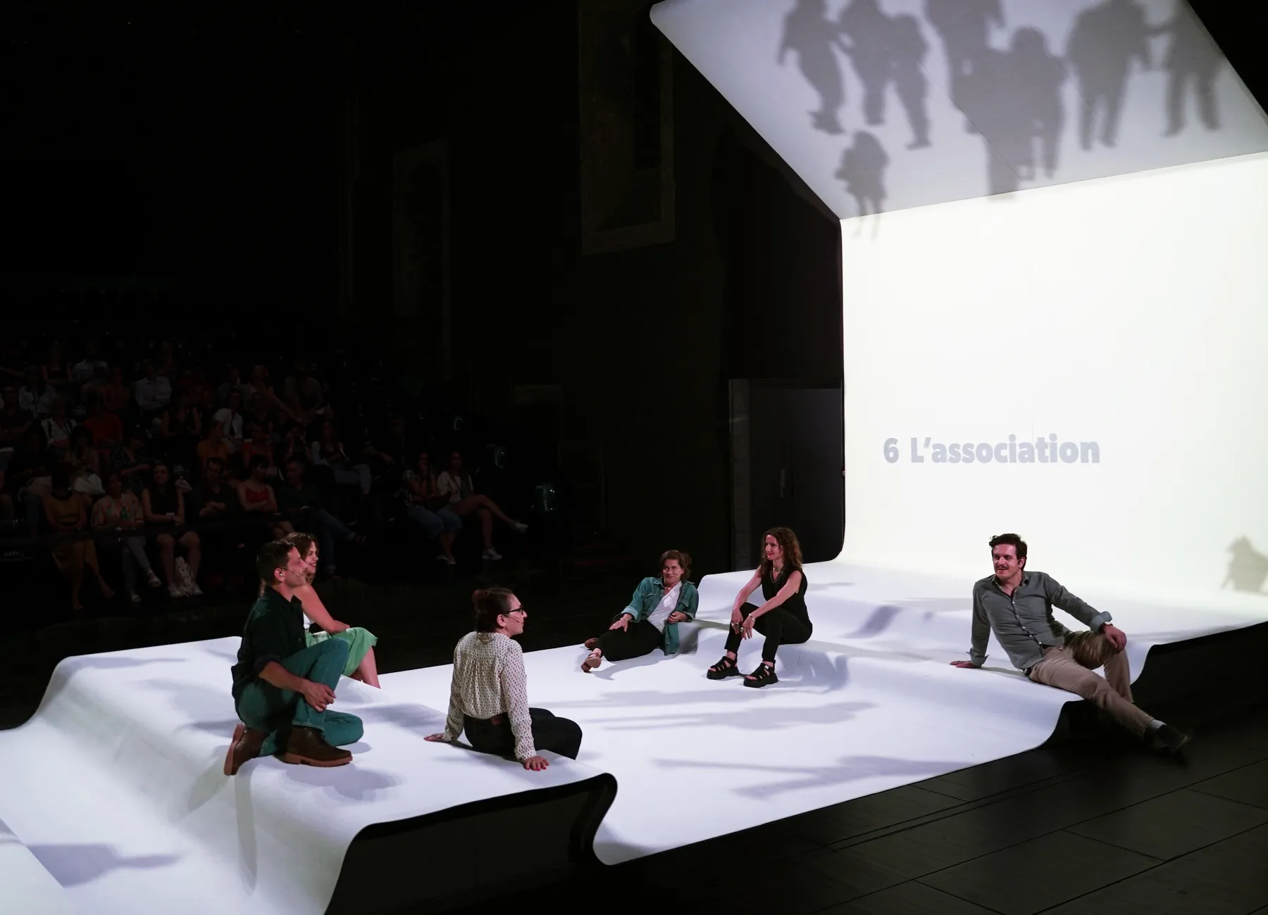 Décor du spectacle Affaires Familiales, tapis et écran blanc, six comédiens assis.