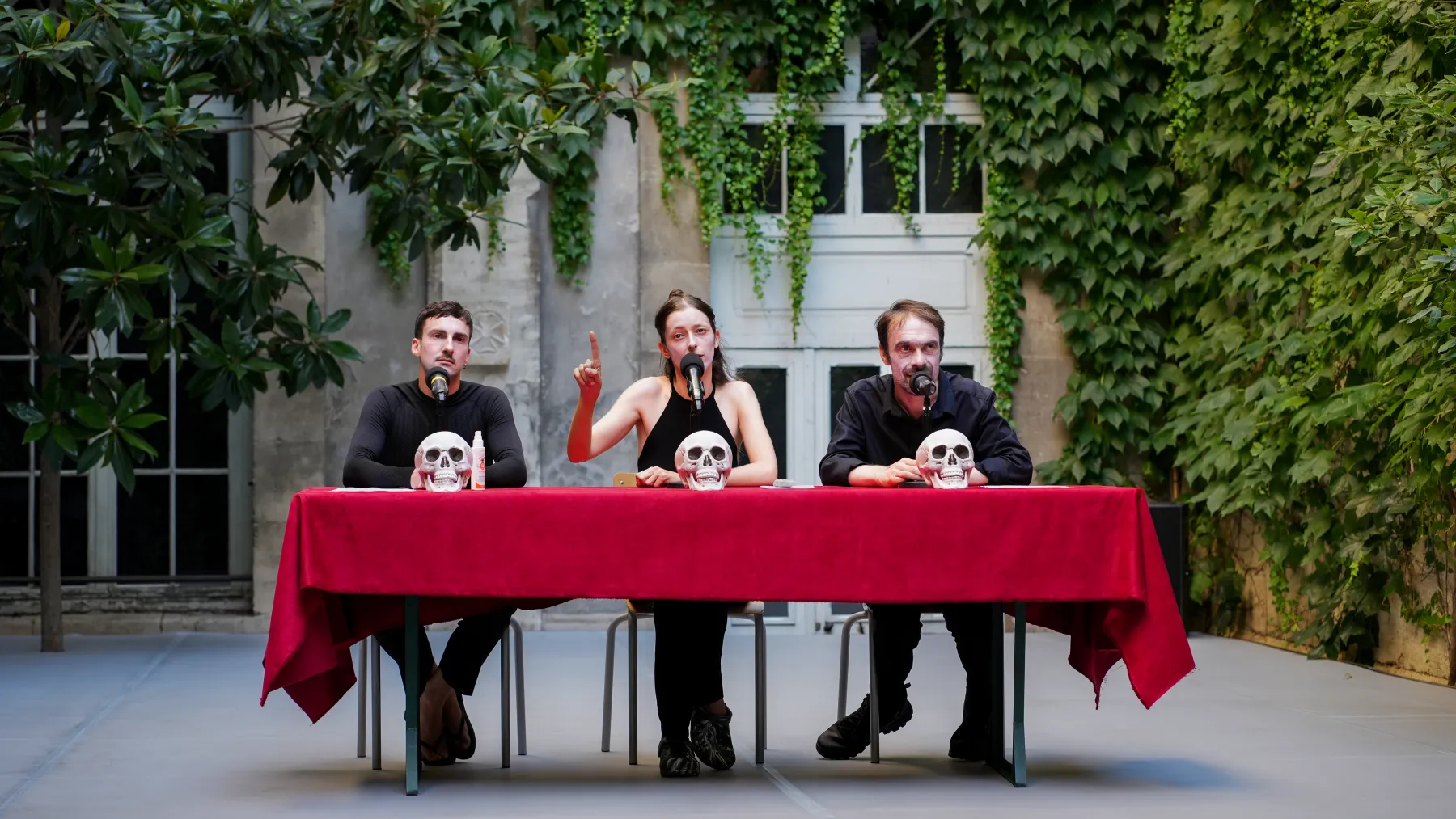 Deux hommes et une femme assis derrière une table rouge, trois crânes humains posés devant eux
