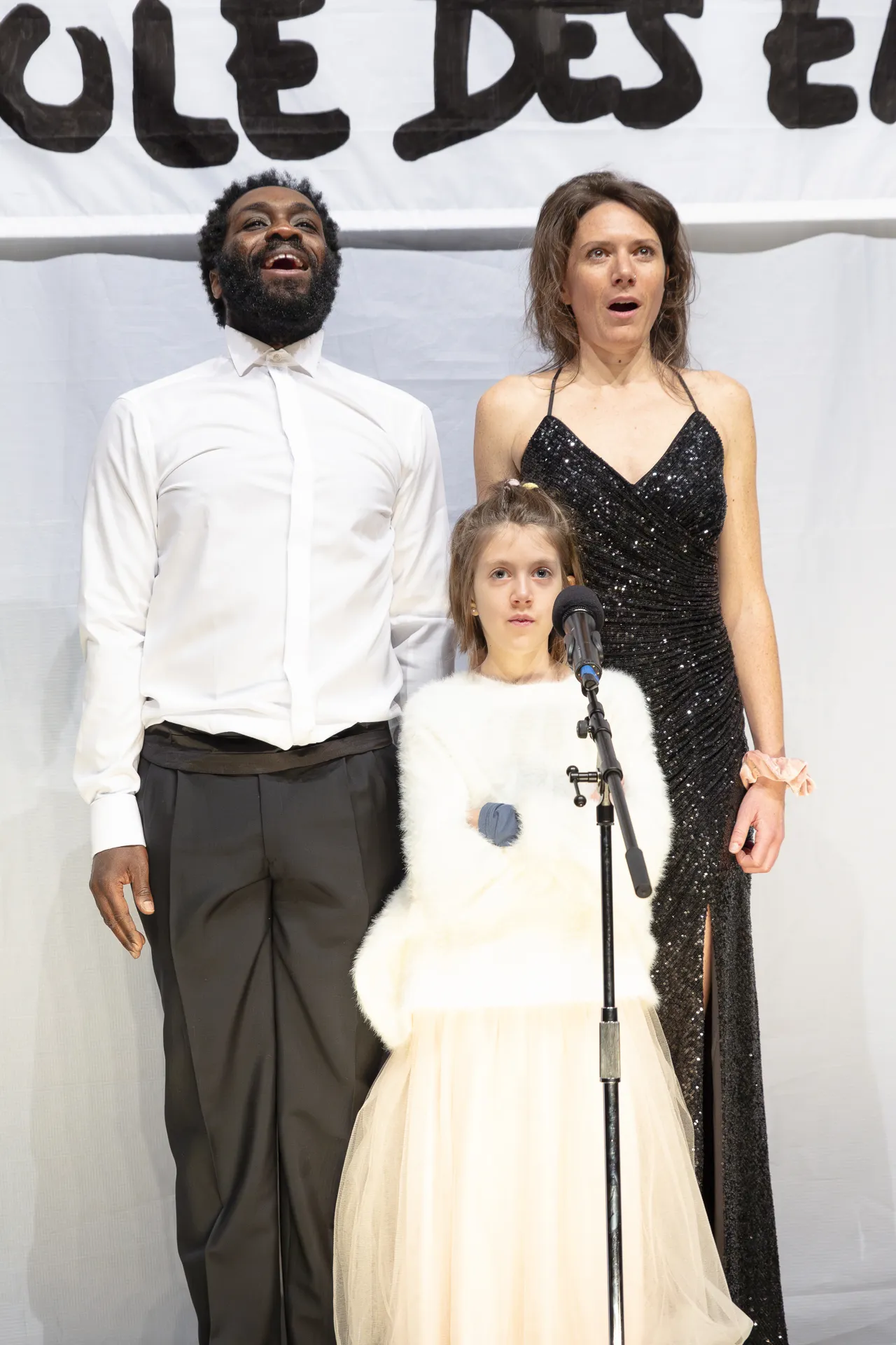 Un homme, une femme et une petite fille devant un micro, les adultes chantent et l'enfant boude